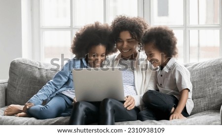 Happy young african American mom relax on sofa at home with two small children use laptop together, smiling biracial mother rest on couch in living room with little kids watch funny video on computer