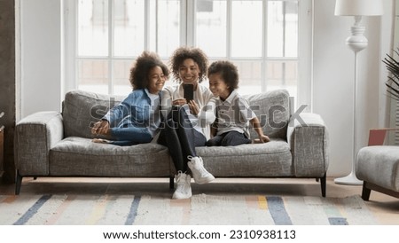 Overjoyed young biracial mother sit relax on couch at home with little children make self-portarit picture on cell, smiling african American mom and two small kids pose for selfie on smartphone