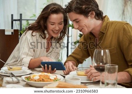 Cheerful young woman showing photo on smartphone to boyfriend when they are eating dinner at home