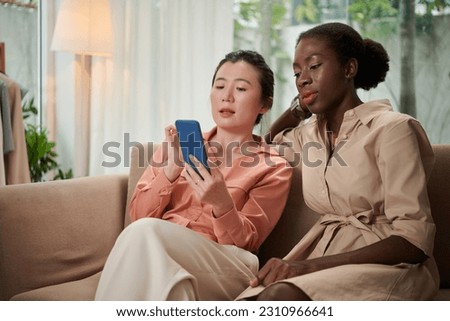 Toung woman sitting on couch with friend and showing her new app with fashion trends