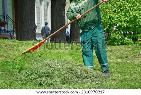 Gardener raking cutted grass with plastic rake, close up. Municipal worker cleaning garbage and mowed grass from green lawn with rake. Man raking mowed grass in the garden. Lawn care service Royalty-Free Stock Photo #2310970917