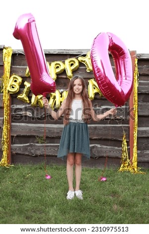 Cute little girl holds two pink foil balloons, digit 10 outdoors. Wooden wall decorated with golden balloons inscription happy birthday.Ten years anniversary celebration event, party at home backyard.