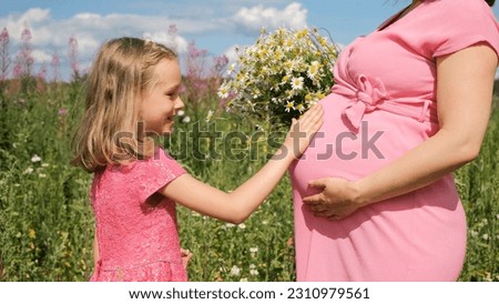 Cute little girl touching belly of her pregnant mother in summer field. Woman holding beautiful bouquet with daisies