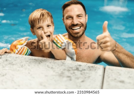Playful father and son  showing thumbs up  while being in swimming pool. There are looking at camera