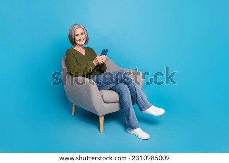 Full length photo of satisfied pleasant woman dressed khaki sweater hold smartphone sit on armchair isolated on blue color background