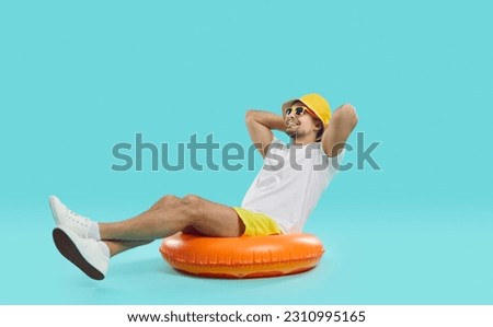 Happy satisfied young man enjoys summer sea vacation sitting on inflatable swimming circle. Joyful guy in summer outfit sits on inflatable circle with his hands behind his head on blue background