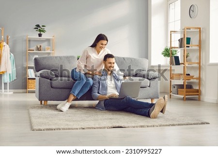Young married couple sitting on sofa in the living room at home using tablet PC for internet and social media. Happy family resting on couch enjoying weekend watching video or talking on video call. Royalty-Free Stock Photo #2310995227