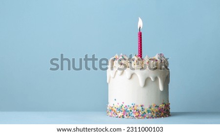 Celebration birthday cake decorated with white drip icing, buttercream frosting swirls, colorful sugar sprinkles and one birthday candle against a plain blue background