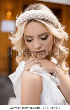 Portrait of adorable young bride with blond hair and pearl headband put hand on shoulder and looking down to reveal wonderful make-up. Expensive looking girl in wedding attire on street of Paris. Royalty-Free Stock Photo #2311012437