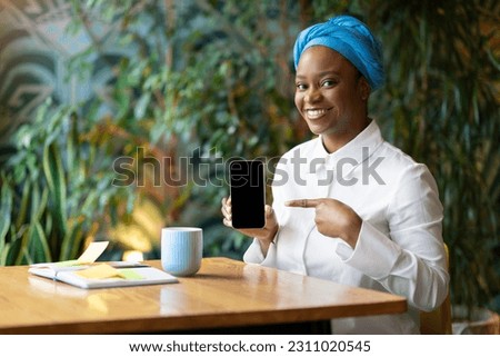 Cheerful positive beautiful young african american woman sitting at cafe, drinking coffee, showing cell phone with white empty screen, recommending nice mobile app for job seeking, mockup