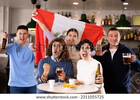 Company of emotional adult young sports fans holding flag of Peru, rejoicing winning match game and drinking beer in bar