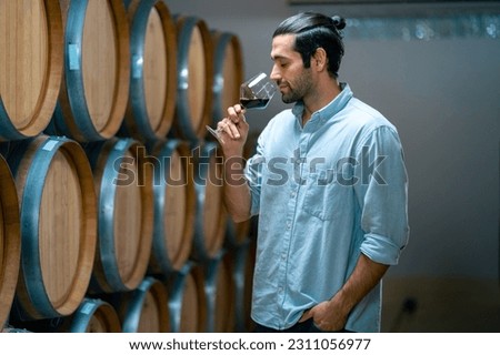 Professional man sommelier testing red wine in wine glass with tasting and smelling at wine cellar with wooden barrel in wine factory. Winery liquor manufacturing industry and winemaker concept. Royalty-Free Stock Photo #2311056977