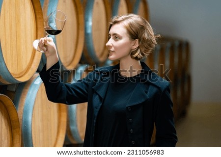 Professional woman sommelier testing red wine in wine glass with tasting and smelling at wine cellar with wooden barrel in wine factory. Winery liquor manufacturing industry and winemaker concept. Royalty-Free Stock Photo #2311056983