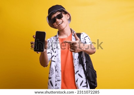 Cheerful Asian tourist traveler man pointing finger to mobile phone screen, recommending online travel agent application over yellow background. 
