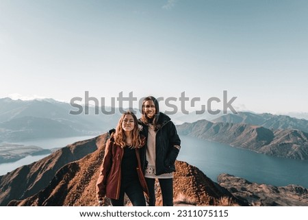 two caucasian girls together one blonde and one brunette looking at camera and smiling happily from the top of the mountain with the big blue lake and mountains on a bright clear day, roys peak, new Royalty-Free Stock Photo #2311075115