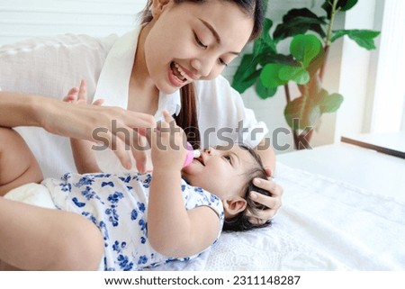 Happy toddle baby infant child lying on white bed while mother holding milk bottle for feeding her daughter kid girl, mom love and take care to new family member, parent look after of child, childhood