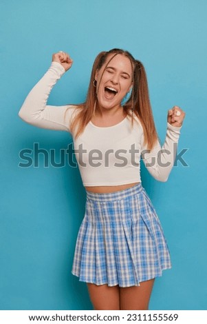 Pretty student girl with two ponytails posing in long sleeve white top and blue checkered miniskirt, dancing with her hands up, happy life concept, copy space