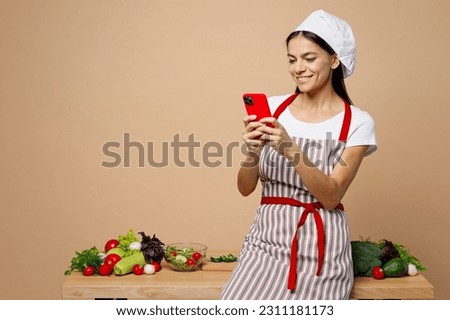 Young smiling housewife housekeeper chef latin woman wear apron toque hat work at table kitchenware use mobile cell phone look recipe isolated on plain pastel light beige background. Process cook food