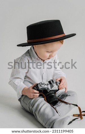 Child in a hat sitting on floor and play with photo camera. Fun kid holds retro vintage camera isolated on white wall. International Photographers Day. Children's studio portrait. Mockup. Close Up. Royalty-Free Stock Photo #2311199405