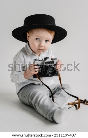 Fun kid holds retro vintage camera isolated on white wall. Child in a hat sitting on floor and play with photo camera. International Photographers Day. Children's studio portrait. Mockup. Close Up.