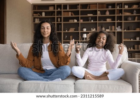 African woman her preschool daughter sit cross-legged on cozy sofa in living room, make mudra gesture do meditation practice at home. Mom teach child good life habit, lifestyle, yoga exercise concept Royalty-Free Stock Photo #2311215589