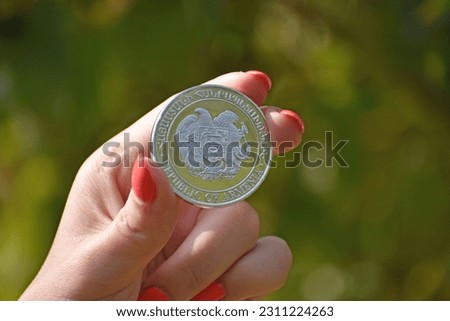 A close up of a woman's hand holding a coin with the Coat of arms of Armenia on it, blurred background
