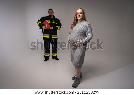 Authentic portrait of a pregnant woman with a firefighter man in uniform, on a light background, full-length photo, place for inscription. Creating and waiting for a young firefighter family