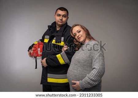 Authentic portrait of a pregnant woman with a firefighter man in uniform, on a light background, full-length photo, place for inscription. Creating and waiting for a young firefighter family