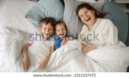 A cheerful young mother in bed with her two sons waving at the camera. This captures the essence of family happiness, spending quality time at home, and joyous moments between parent and child