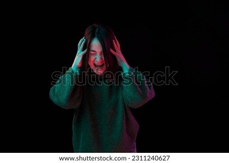 Anger, irritation, stress. Young girl in cozy sweater emotionally posing screaming over dark background in neon light. Concept of emotions, facial expression, youth, inspiration, sales, ad