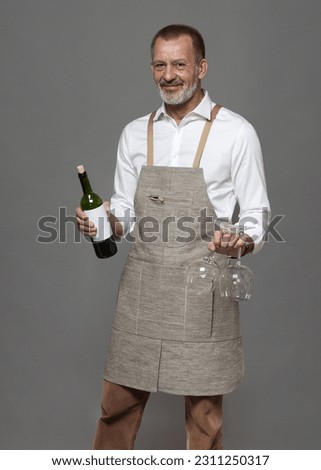 Respectable man in a linen apron holds wine glasses and a bottle of wine. Sommelier or waiter in joy and hospitality. Royalty-Free Stock Photo #2311250317