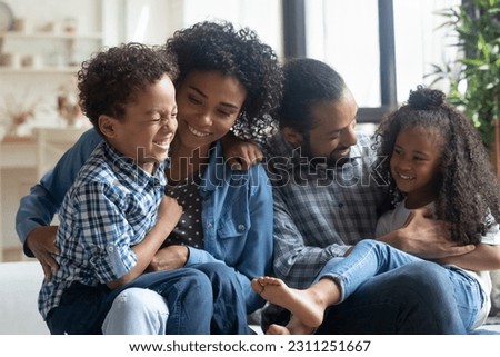 Happy African American parents with two kids having fun, sitting on couch, smiling mother and father with laughing little son and daughter hugging, tickling, enjoying leisure time weekend at home