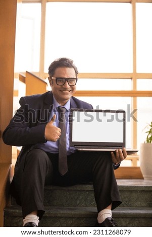 Indian corporate man showing thumps up with laptop empty screen at office.