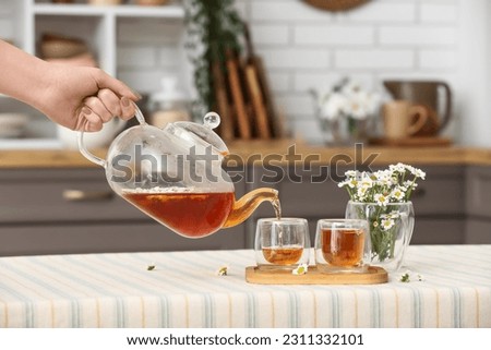 Female hand pouring natural chamomile tea from teapot into cup on table in kitchen Royalty-Free Stock Photo #2311332101