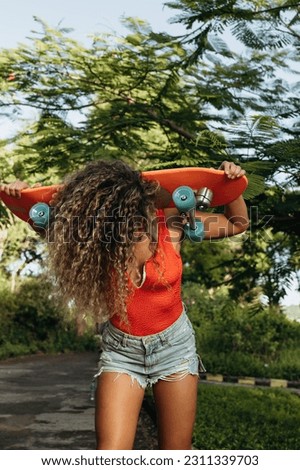 Young girl's face is covered with long wavy hair, dressed in stylish clothes, holding a skateboard over her head in a park on a bright sunny day