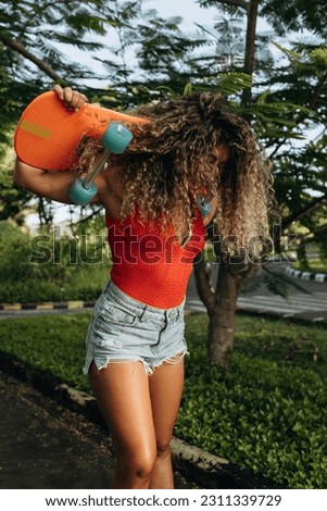 Portrait of a young happy girl with long wavy hair in nature laughing and holding a skateboard behind her head in a skate park. Active lifestyle