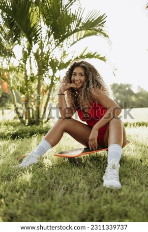 Happy Asian girl with long wavy hair sits on a skateboard in the park, laughs and smiles. Extreme sport, healthy lifestyle.