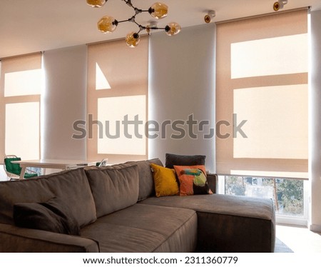 Motorized roller blinds. A sofa with colorful pillows in the room near windows with sunscreen curtain. Automatic roller shades on full height windows in the interior. Sunny day. Selective fokus. Royalty-Free Stock Photo #2311360779