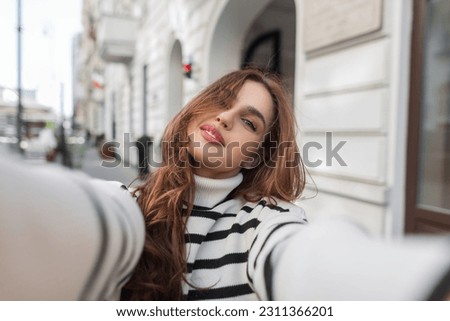 Beautiful fashion young woman in a stylish striped sweater walks in the city and takes a selfie photo on a smartphone. Woman travels and takes a photo