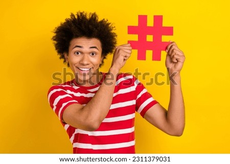 Photo of cheerful positive guy dressed red t-shirt smiling rising hash tag card isolated yellow color background