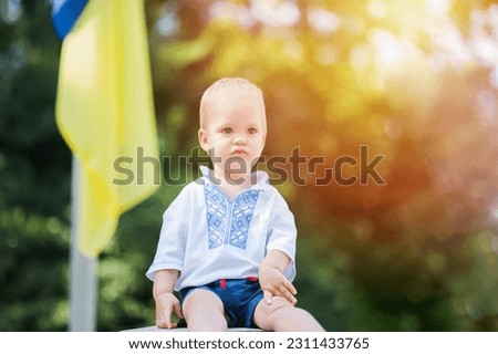 Toddler boy in Ukrainian vyshyvanka on background of Ukrainian yellow-blue flag in summer.