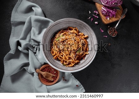 Overhead, horizontal view of stir-fried udon noodles with chicken and vegetables, beautifully arranged on a black background, with spices and a napkin lending character. Royalty-Free Stock Photo #2311460997