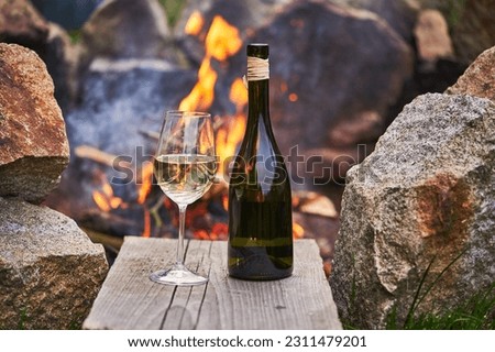 Romantic picture of the glass of white wine and rustic style bottle from small family winery in front of burning stone campfire during the summer sunset in the south moravian region of Czech Republic. Royalty-Free Stock Photo #2311479201