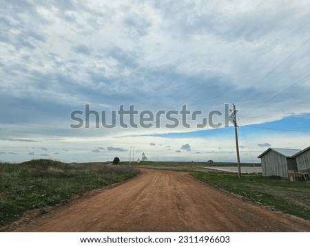 A red dirt road under a cloudy afternoon sky with a lighthouse in the far distance. Royalty-Free Stock Photo #2311496603