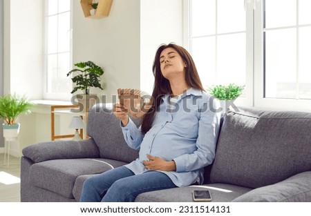 Pregnant mother woman in third trimester expects baby, suffers from extreme summer heat and hot flush hormone change symptoms, sits on sofa in too stuffy overheated room, and refreshes with hand fan Royalty-Free Stock Photo #2311514131