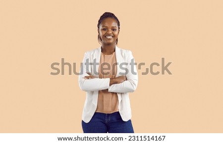 Studio shot of happy confident young black businesswoman. Portrait of smiling good looking African American business lady in white jacket standing with arms folded isolated on solid beige background