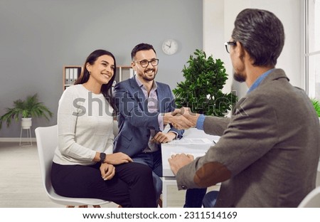 Happy family couple making real estate purchase agreement. Young man smiling and shaking hands with realtor or estate agent while sitting together with his wife at desk in estate agency office