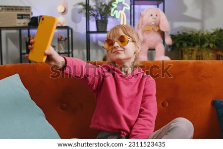 Young blonde child kid girl blogger taking selfie on smartphone, communicating video call online with social media subscribers. Female teen toddler recording video story at home in room sits on sofa
