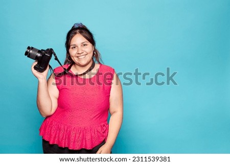 Young curvy latina woman smiling holding reflex camera in hand. Indoor studio shot isolated on blue background. Copy space. Royalty-Free Stock Photo #2311539381