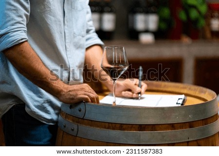 Professional man sommelier testing red wine in wine glass with tasting and smelling at wine cellar with wooden barrel in wine factory. Winery liquor manufacturing industry and winemaker concept. Royalty-Free Stock Photo #2311587383